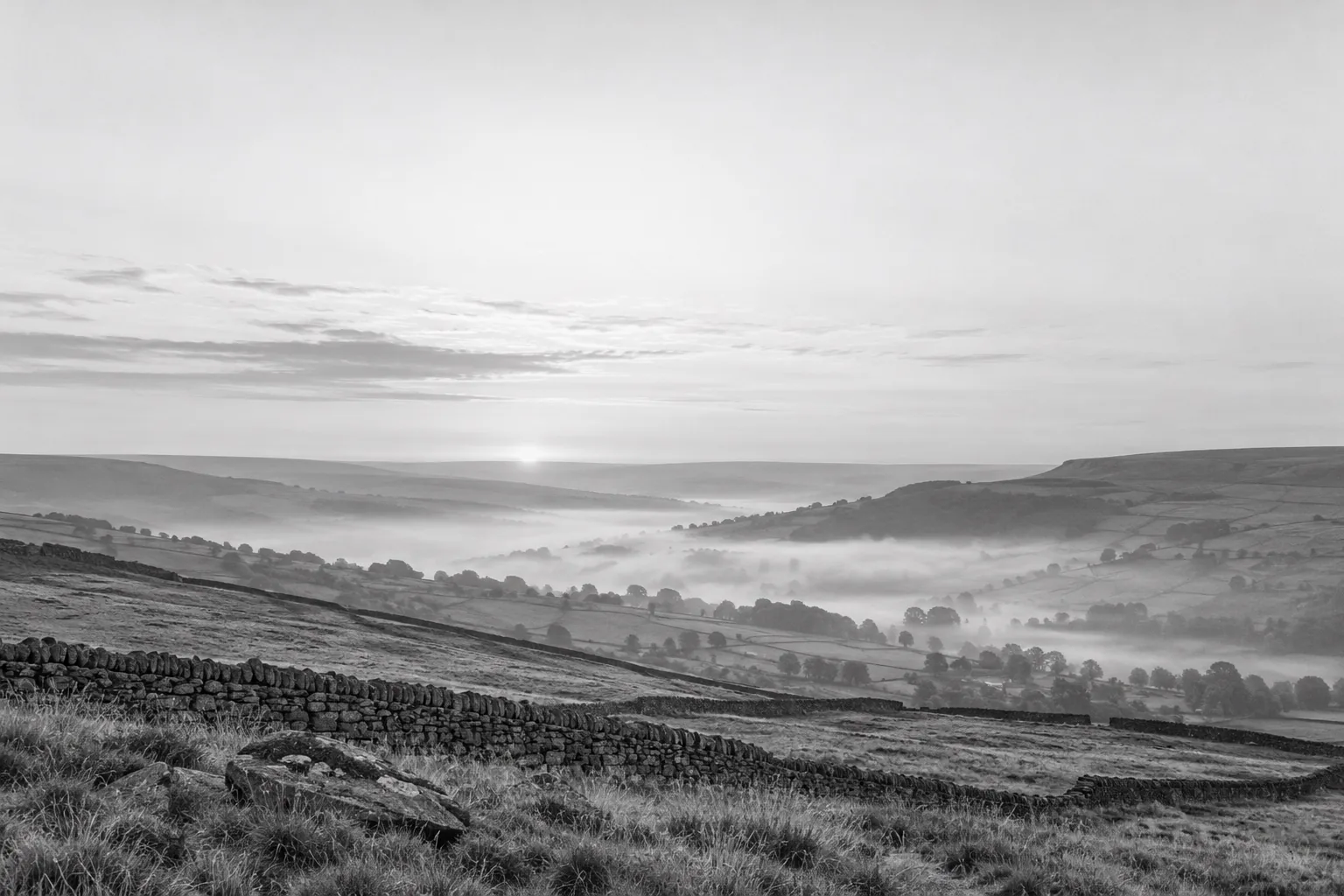 Pennine moorland at dawn in black and white, dry-stone walls running down to a valley filled with mist under a pale sky